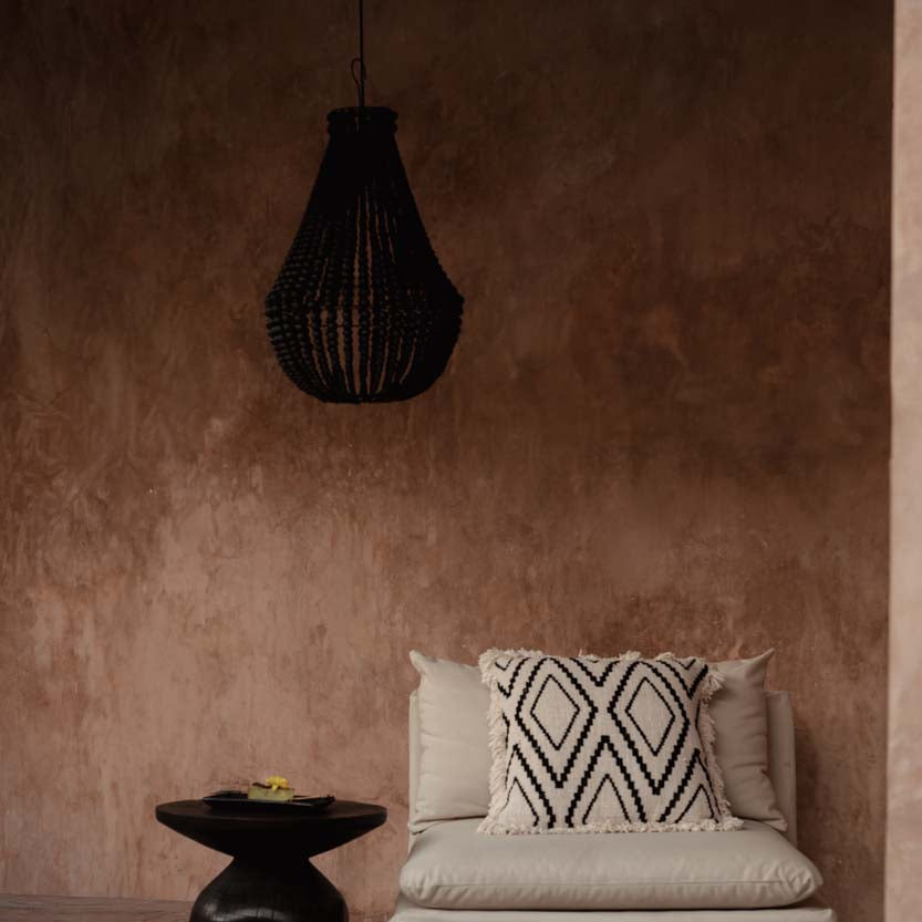 Beige armchair with patterned pillow and black side table against a brown wall with black chandelier.
