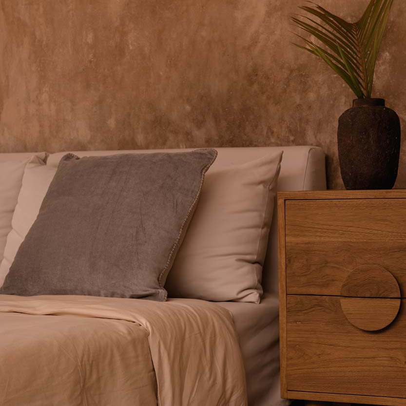 Bedroom with beige bedding, wooden nightstand, and decorative plant against a brown wall.