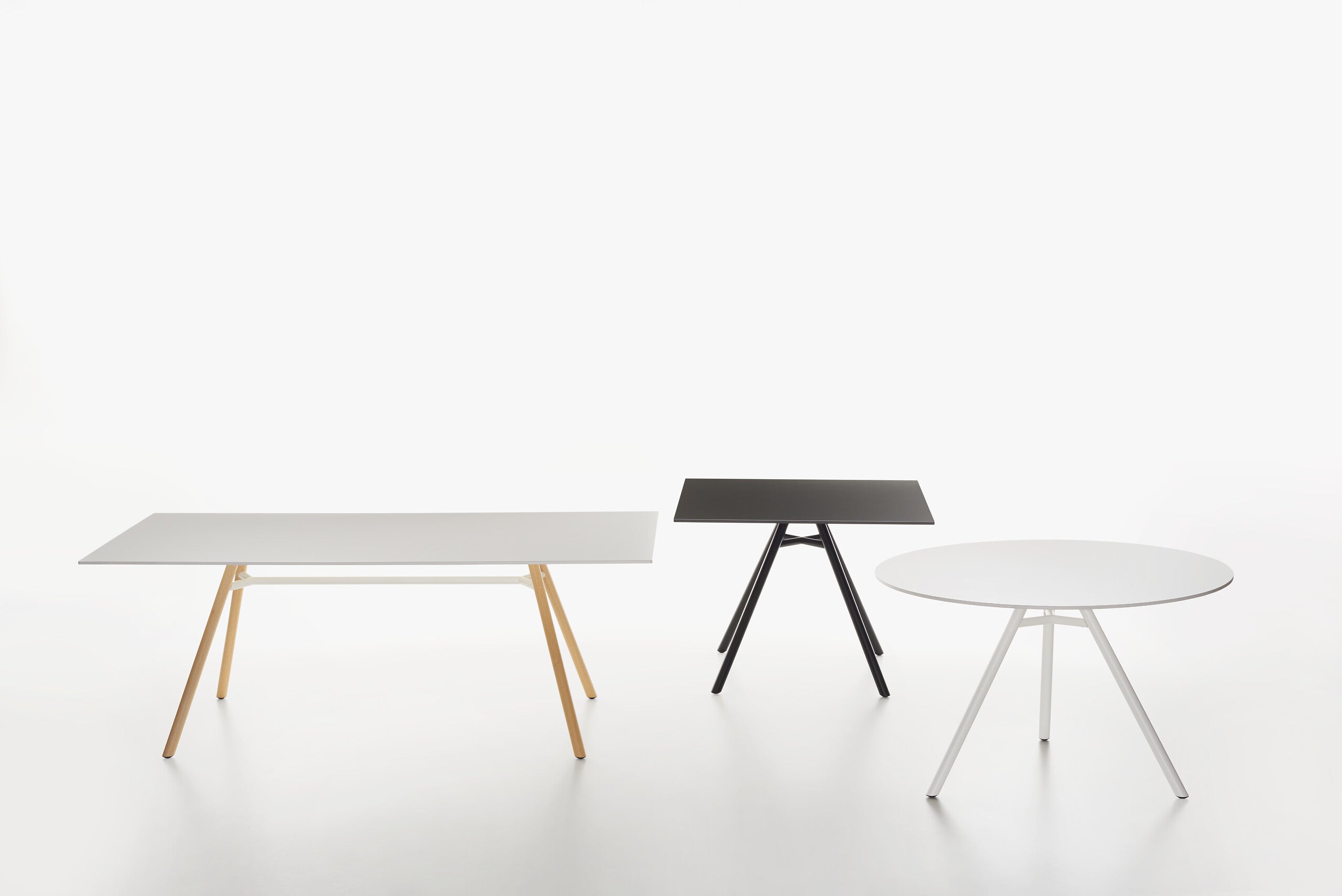 Three modern minimalist MART tables on a seamless white studio background: a  rectangular table with a white top, a square table with a black top and black legs, and a round white table with white legs.